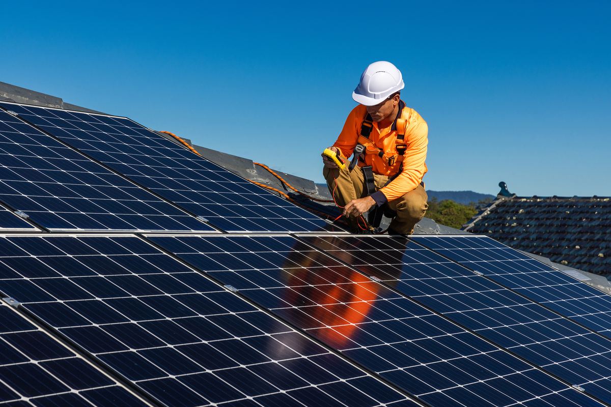 An installer in a hard hat and hi-vis kneeling on a large roof-mounted solar array.