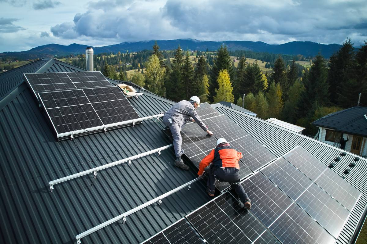 Two installers fixing solar panels to mounting rails on a long-run metal roof, with forest and hills behind.