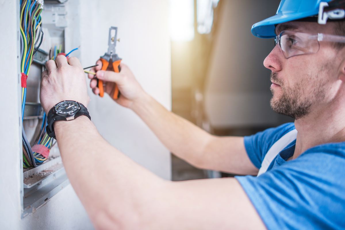 An electrician in a hard hat and safety glasses wiring a wall box, pliers in hand.