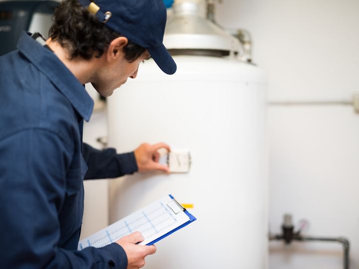 A technician in overalls and a cap adjusting the control on a white hot water cylinder, clipboard in hand.