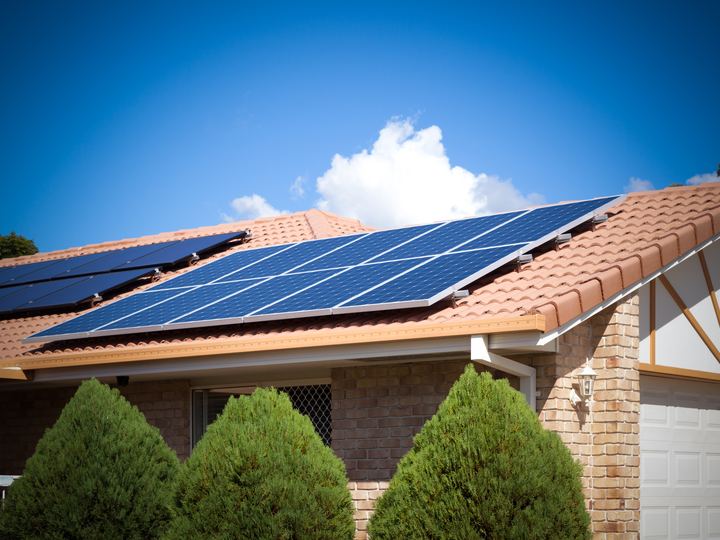 Solar panels on the tiled roof of a single-storey brick house, conifers in front.